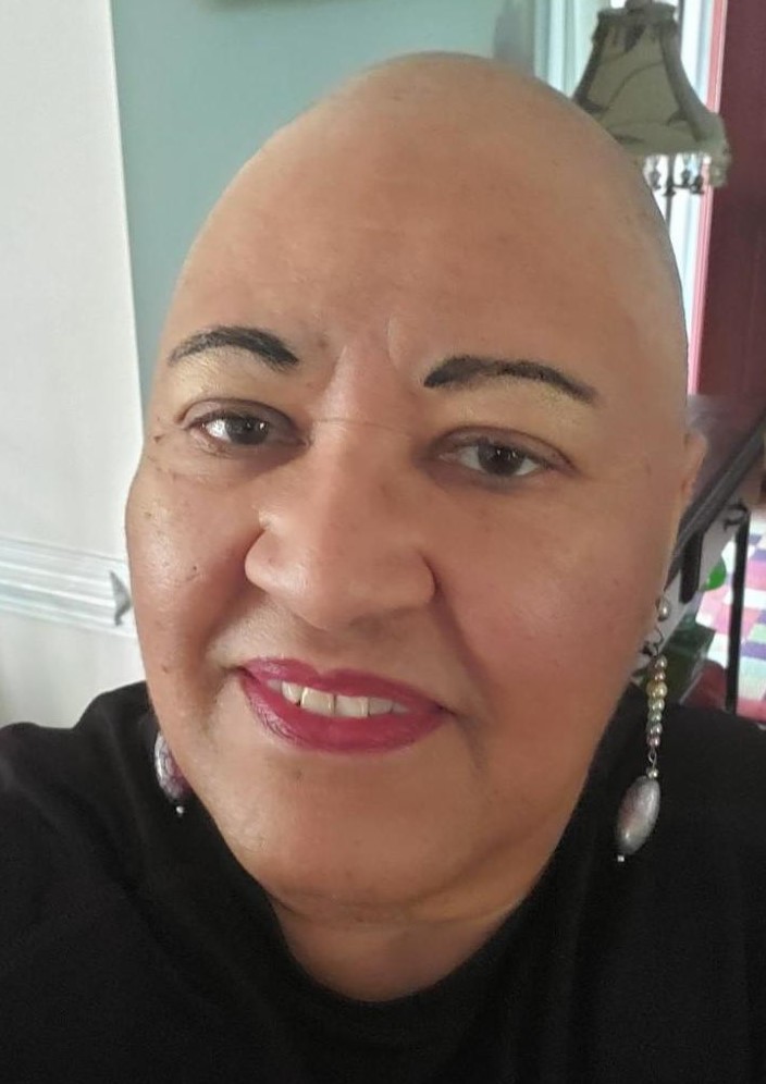 Linda Mason, author, a light skinned Black woman with brown eyes. She is wearing light gold eye shadow, darkened eye brows, and deep pink lipstick along with dangling earrings. She is smiling and has no hair on the top of her head.
